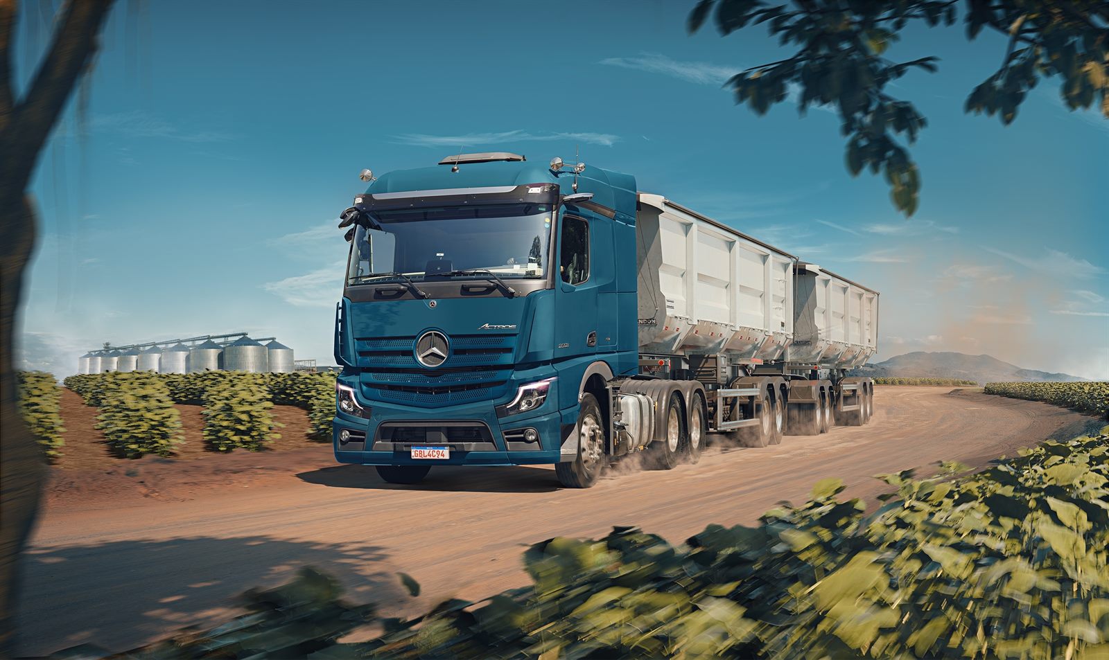 Caminhão Mercedes-Benz Actros com bitrem carregado em estrada de terra entre lavouras, com silos de armazenagem ao fundo.